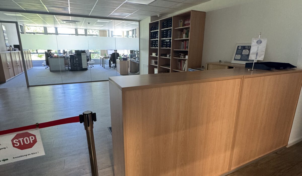 Empfangsbereich im Büro mit hölzerner Theke, Absperrband mit Stop-Schild und Blick durch Glaswand in den Arbeitsbereich.