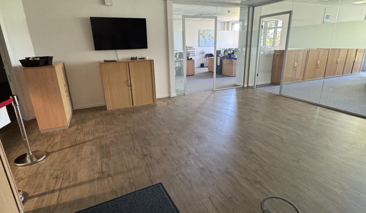 Moderner Bürobereich mit Glaswänden, Vinylboden und Blick in helle Büroräume mit Schreibtischen und Schränken.