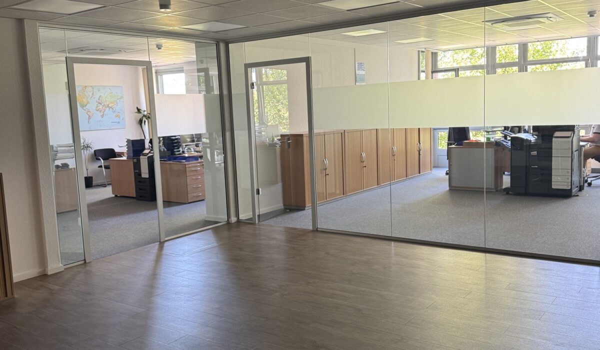 Moderner Aufenthaltsraum im Büro mit Holzschränken, dunkler Schmutzfangmatte und Glasabtrennung zum Büro.