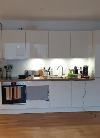 Moderne weiße Einbauküche mit Holz-Arbeitsplatte, beleuchteter Nische, Backofen und Ceranfeld.